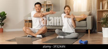 Uomo e donna asiatici felici che si allungano insieme su un tappetino da yoga a casa, concetto di perdita di peso Foto Stock