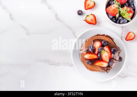 Pila di pancake al cioccolato con una ricca crema di pasta al cioccolato e frutti di bosco freschi - fragole, mirtilli. Colazione dolce e gustosa Foto Stock