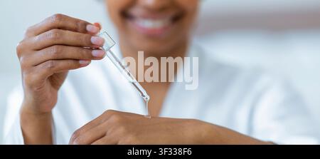Donna nera sorridente che applica siero sulla pelle con un contagocce a casa, routine di cura della pelle Foto Stock