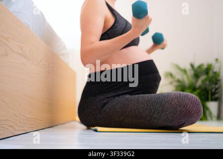 Una donna incinta si siede su un tappeto da yoga tenendo un manubrio, praticando yoga prenatale sul pavimento a casa. La madre e atleta mostra un benessere calmo e consapevole e una forma fisica pacifica ed equilibrata in una palestra domestica. Foto Stock