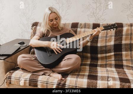 Una donna matura è seduta a gambe incrociate su un divano e si esercita a suonare una chitarra acustica nera accanto a un laptop. Foto Stock