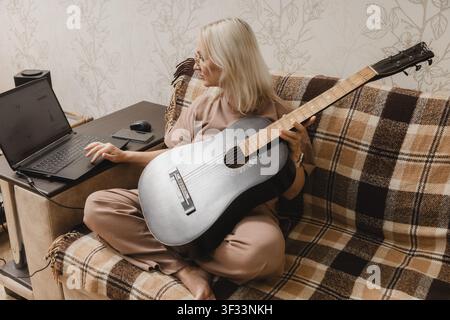 Una donna matura è seduta su un divano con una chitarra acustica e usa un laptop nel suo soggiorno. Foto Stock