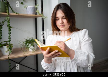 Giovane donna che scrive in un quaderno giallo mentre studia a casa vicino a una libreria con piante. Educazione e sviluppo personale in un ambiente domestico accogliente Foto Stock