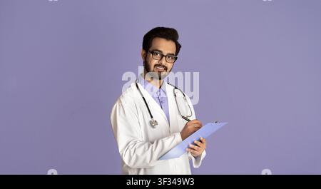 Ritratto di giovane medico indiano maschio che scrive sulla clipboard sopra sfondo lilla Foto Stock
