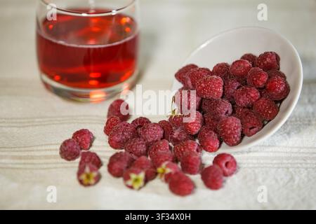 Un primo piano orizzontale di lamponi rossi maturi che fuoriescono da una ciotola di ceramica bianca su una tovaglia di lino. Un bicchiere di succo rosso è leggermente sfocato Foto Stock