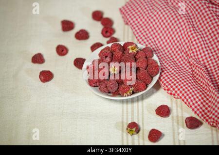 Una fotografia ad angolo alto di una piccola ciotola bianca piena di lamponi rossi maturi su una tovaglia di lino chiara. Diversi frutti sono sparsi intorno, e Foto Stock