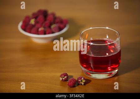 Un primo piano orizzontale di un bicchiere trasparente pieno di una vivace bevanda rossa alla fragola su una superficie in legno liscio. Tre fragole fresche giacciono i Foto Stock