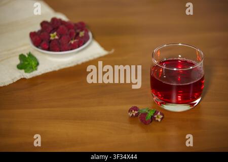 Un colpo orizzontale ad angolo alto di un bicchiere con succo di lamponi rosso su un tavolo di legno. sullo sfondo, una ciotola bianca piena di lamponi si trova su un Foto Stock
