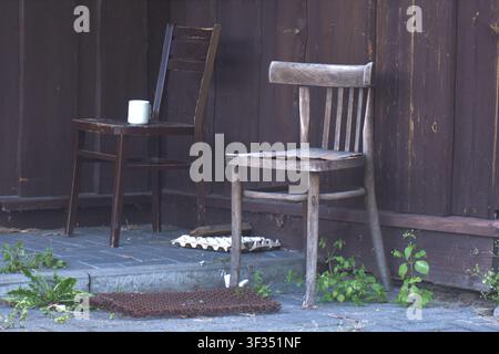 Due vecchie sedie di legno appoggiate contro un muro rustico con una tazza dimenticata, che evocano nostalgia, solitudine tranquilla e una vita lenta e semplice. Foto Stock