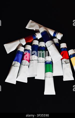 Tubi di vernice colorati sparsi su uno sfondo nero scuro. Foto di alta qualità Primo piano di tubi di vernice acrilica o a olio chiusi disposti a forma di ventaglio sparso su uno sfondo nero solido. Lo sfondo scuro crea un forte contrasto, evidenziando i colori vivaci sulle etichette dei tubi. Perfetto per concetti di materiale artistico, studi creativi, hobby della pittura e sfondi di design artistico. Foto Stock