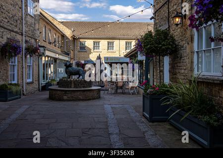 Affascinante cortile lastricato nella storica città inglese di Cirencester con edifici in pietra, vasi per fiori e una fontana a forma di statua di pecora Foto Stock