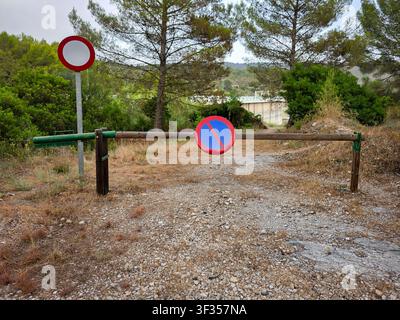 Una barriera in legno è chiusa su un sentiero di ghiaia che conduce a una diga. Accanto alla barriera sono visibili cartelli „Divieto di sosta“ e „Divieto di accesso“. Alberi e cespugli surfano Foto Stock