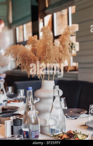 Il tavolo da pranzo contiene piatti con cibo. Un vaso bianco contiene erba della pampa. Sono disposte bottiglie d'acqua e bicchieri da bere. Lo sfondo mostra un ristorante Foto Stock