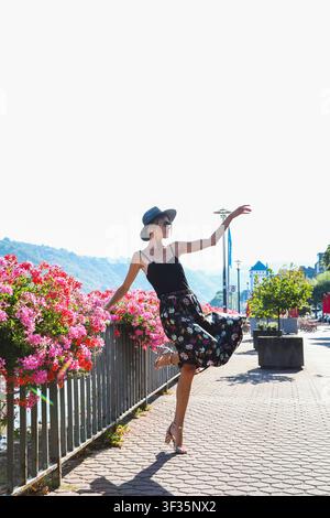 Donna elegante in una gonna di seta floreale fluida e una canottiera, in piedi accanto a una recinzione decorata con gerani in fiore su una passeggiata europea soleggiata. Foto Stock