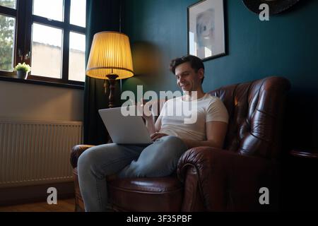 Giovane uomo sorridente gesticola durante una videochiamata su un laptop mentre è seduto su una poltrona di pelle a Praga, Repubblica Ceca. Foto Stock