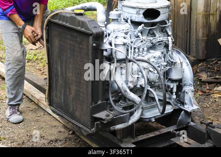 Un motore diesel su telaio metallico in riparazione all'aperto da parte di un meccanico, radiatore e tubi visibili, ambiente di officina ruvido con strumenti e componenti usurati Foto Stock