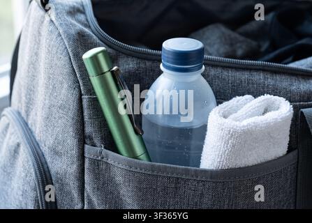 Primo piano di una borsa sportiva in tessuto grigio. La tasca laterale contiene una bottiglia d'acqua raffreddata con condensa, una torcia in metallo verde e un asciugamano bianco arrotolato Foto Stock
