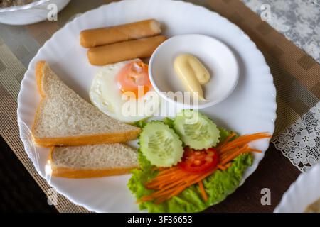 Un uovo fritto, salsicce, triangoli di pane, maionese e insalata fresca disposti su un piatto bianco per una colazione accogliente Foto Stock