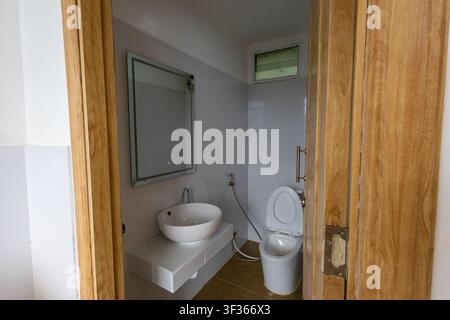 Un interno di bagno moderno con lavandino in ceramica bianca, specchio da parete e toilette, telaio della porta in legno naturale e luce naturale soffusa che crea un'atmosfera calma e pulita Foto Stock