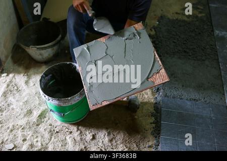 Una piastrella spalmata con malta bagnata sulla cazzuola per l'installazione del pavimento, operaio edile che applica adesivo e liscia il cemento con un secchio e delle piastrelle Foto Stock