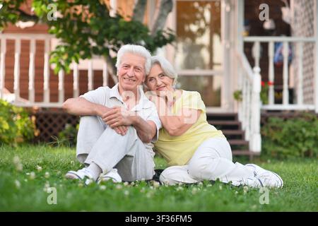 Anziano seduto sull'erba davanti alla casa, rilassato e soddisfatto, a livello del suolo, leggermente rialzato, materiali in cemento e legno, verde e bianco Foto Stock