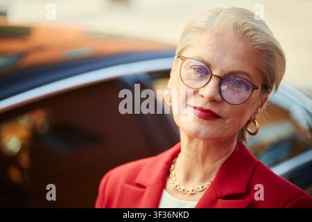 Portratto di una donna d'affari caucasica anziana che guarda verso la telecamera vicino a un'auto, che trasmette sicurezza ed esperienza esecutive. Ideale per leadership, crescita professionale, marketing di servizi di lusso Foto Stock