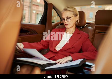 Donna d'affari caucasica di mezza età che esamina documenti sul sedile posteriore dell'auto, preparandosi per una riunione durante il tragitto. Mobilità aziendale, viaggi esecutivi, uso per campagne di leadership Foto Stock