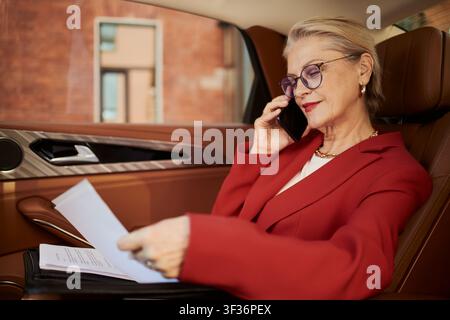 Donna d'affari caucasica anziana che parla al smartphone e rivede documenti mentre è in macchina, gestendo il flusso di lavoro mobile. Ideale per viaggi esecutivi, leadership, marketing della produttività Foto Stock