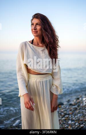 Giovane donna in piedi su una spiaggia di ciottoli durante l'ora dorata, indossa un abito estivo leggero e guarda verso l'orizzonte Foto Stock