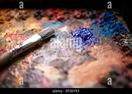 Primo piano di un pennello usato appoggiato su una tavolozza artistica ruvida con pitture a olio miste e colori vivaci Foto Stock