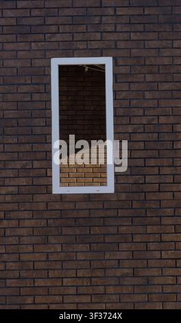 Astrazione architettonica minimalista con una cornice bianca della finestra su un muro di mattoni marroni che riflette un edificio di mattoni opposto nella luce del sole e nell'ombra. Foto Stock
