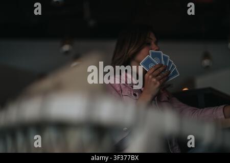 Donna che tiene carte da gioco in mano mentre è seduta al chiuso in una scena incontroluce di atmosfera suggestiva Foto Stock