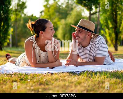 Una coppia multirazziale si gode un tranquillo picnic in un rigoglioso parco verde, sdraiata su una coperta e sorridendosi calorosamente. Foto Stock
