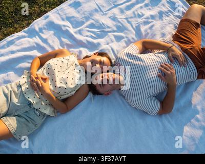 Una coppia sorridente giace fianco a fianco su una coperta da picnic, godendosi un pomeriggio tranquillo in un parco illuminato dal sole. Foto Stock