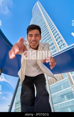 Giovane uomo sorridente che tende la mano verso la fotocamera, in piedi davanti a un grattacielo imponente e un cielo azzurro in una zona urbana moderna Foto Stock