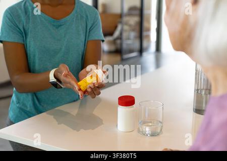Diversa donna assistente e anziana che versa pillole nella mano a coppa da una bottiglia arancione a casa Foto Stock