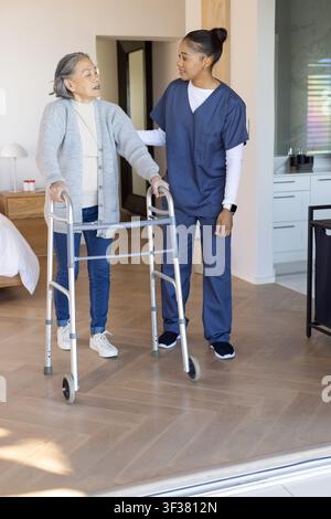 Donna asiatica anziana che utilizza un deambulatore a quattro ruote mentre un'addetta all'assistenza in camice blu marina offre supporto in camera Foto Stock