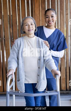 Anziana asiatica in piedi con deambulatore, badante afroamericana in divisa che la sostiene sul portico. Anziana, infermiera, mobilità, assistenza, all'aperto, pat Foto Stock