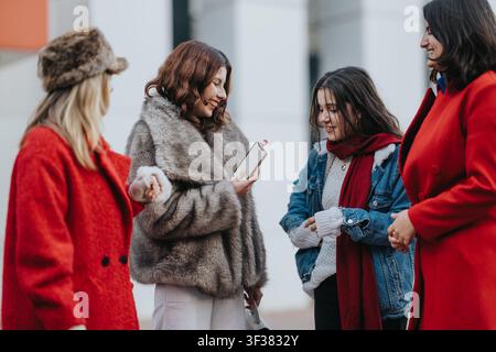 Quattro amiche che chiacchierano e ridono all'aperto con giacche invernali mentre guardano uno smartphone Foto Stock
