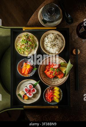 Vista dall'alto di un set per pranzo giapponese su un vassoio nero che include sashimi di salmone, rotoli di sushi maki, ciotola di riso, pollo croccante e insalata fresca. Foto Stock