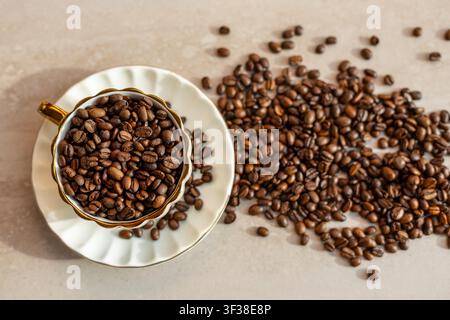 Caffè tostato in tazza e piattino decorativi. Vista dall'alto di chicchi di caffè tostati che riempiono una tazza bianca decorativa e sparsi su un beige Foto Stock