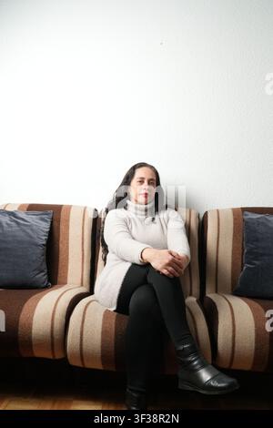Donna seduta su un divano a righe, mani giunte. Indossa un dolcevita e stivali neri. Cuscini scuri la fiancheggiano su entrambi i lati. Il muro dietro di lei è semplice e Foto Stock
