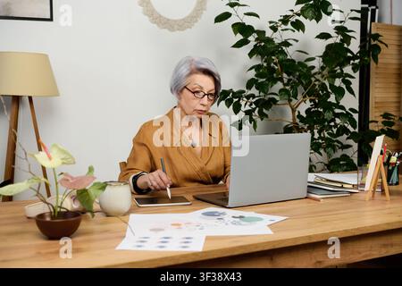 Donna anziana con capelli grigi si impegna attivamente nel lavoro a distanza dal suo ufficio di casa Foto Stock