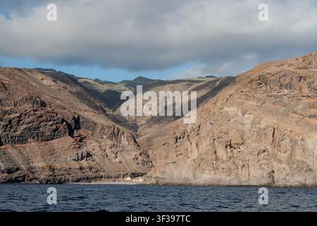Montagne vulcaniche aspre e una valle ripida che incontra l'Atlantico blu profondo sopra un minuscolo insediamento sulla costa di La Gomera, Isole Canarie, Spagna. Foto Stock