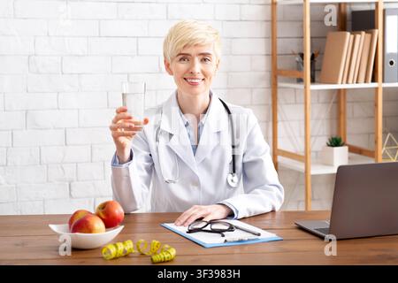 Il nutrizionista del medico felice guarda la macchina fotografica, tiene il vetro dell'acqua, siede al tavolo con le mele sul piatto Foto Stock