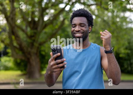 Giovane uomo entusiasta con un grande sorriso e il pugno stretto che prova gioia e realizzazione mentre legge buone notizie sul suo telefono cellulare all'aperto in un ambiente di parco Foto Stock