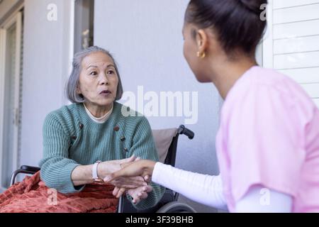 Donna anziana asiatica seduta su una sedia a rotelle che tiene la mano di un'infermiera in uniforme rosa sul portico Foto Stock