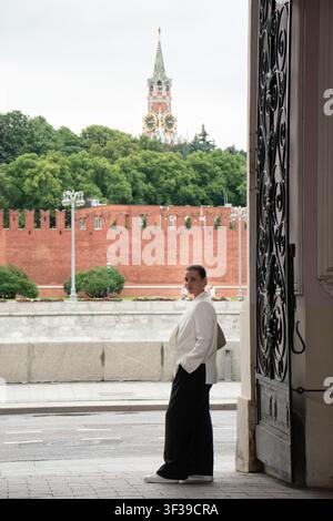 Donna elegante in piedi sotto un arco storico che si affaccia sul muro del Cremlino e sulla torre Spasskaya nel centro di Mosca in una giornata nuvolosa. Stile di vita urbano, viaggio Foto Stock