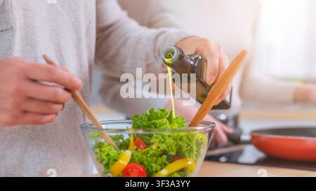Mani irriconoscibili che versano olio d'oliva in un'insalata fresca in una cucina moderna Foto Stock