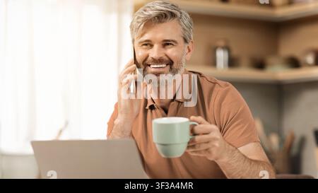 Uomo caucasico maturo sorridente che parla al telefono con una tazza di caffè a casa, concetto di ufficio domestico Foto Stock